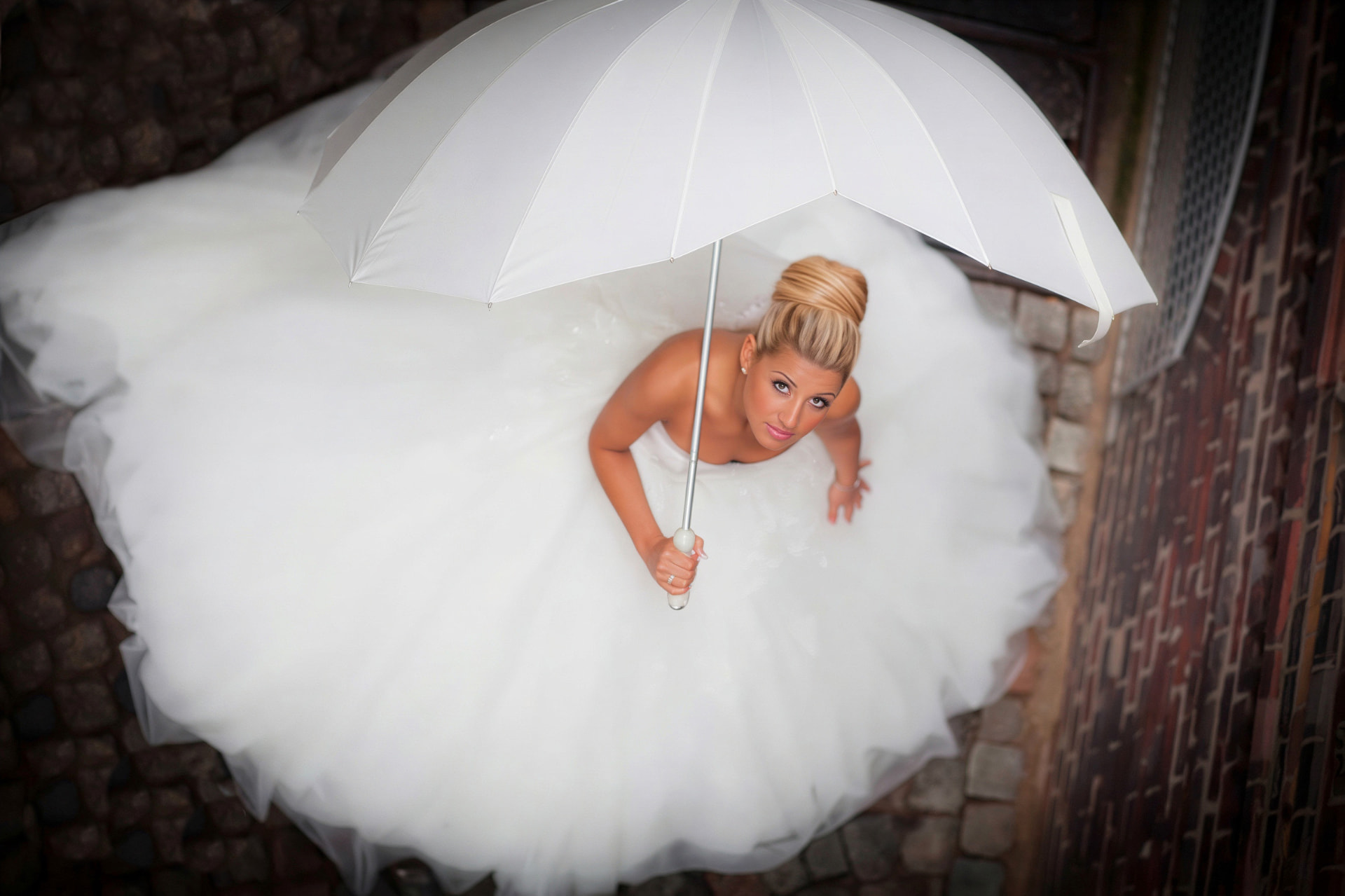 Braut mit weißem Schirm im ausgebreiteten Tüllkleid auf Kopfsteinpflaster, von oben fotografiert