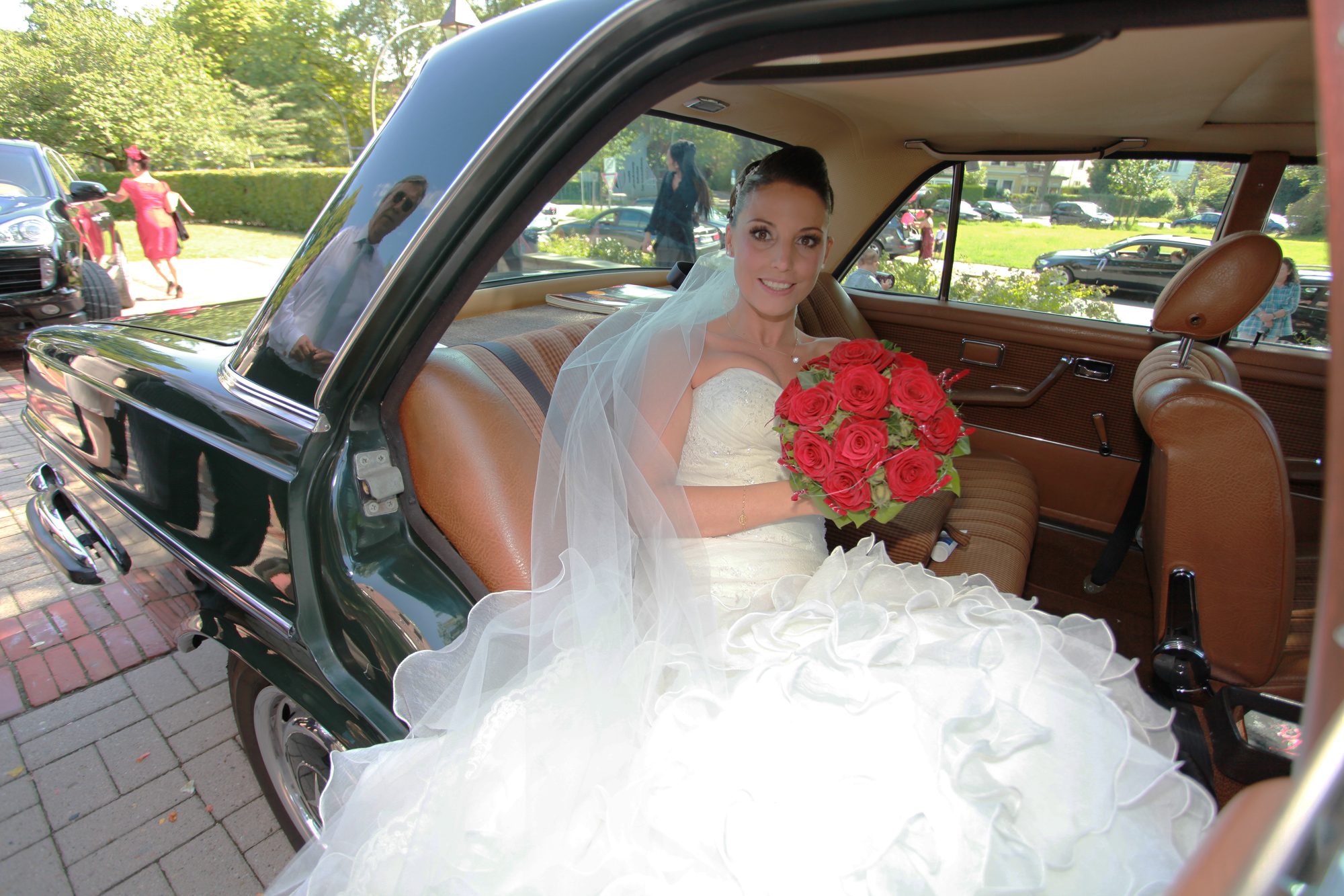 Lächelnde Braut mit rotem Rosenstrauß sitzt im Fond eines dunkelgrünen Oldtimers, Hochzeitsfotograf Hamburg