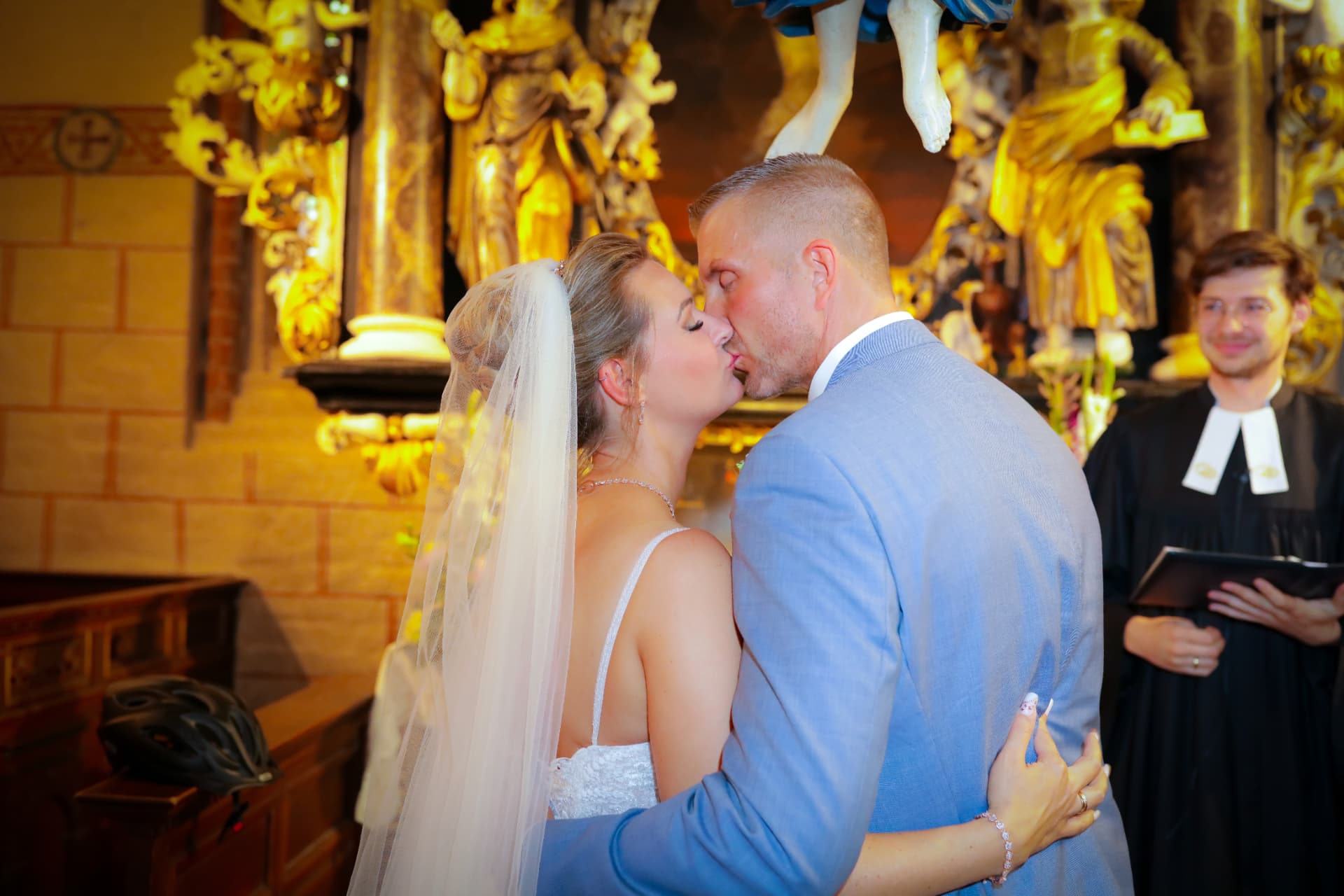 Brautpaar küsst sich vor dem goldenen Barockaltar, der Pastor lächelt im Hintergrund